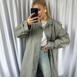 May include: A long, gray trench coat with a belt and button closure. The coat is worn over a white t-shirt and light blue jeans.