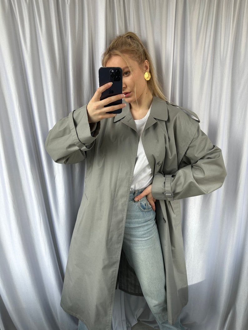 May include: A long, gray trench coat with a belt and button closure. The coat is worn over a white t-shirt and light blue jeans.