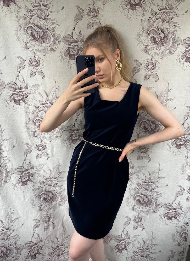 Pu&ograve; includere: Un abito in velluto blu navy senza maniche, con scollo quadrato e cintura a catena dorata.