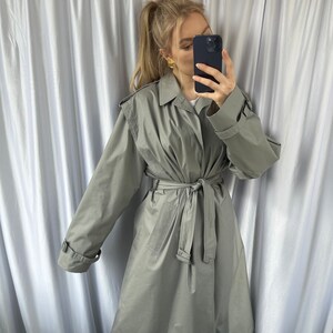 May include: A light gray trench coat with a belt and long sleeves. The coat has a classic design and is made of a lightweight fabric.