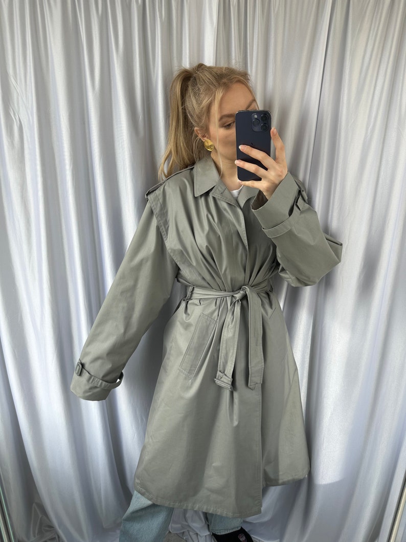 May include: A light gray trench coat with a belt and long sleeves. The coat has a classic design and is made of a lightweight fabric.
