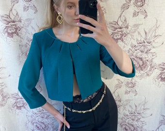 Pode incluir: Um blazer cropped turquesa com decote em V e mangas três quartos. O blazer é usado sobre um top de renda preto e calças pretas com um cinto de corrente dourado.