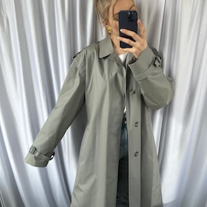 May include: A long, gray trench coat with a button closure and a belt. The coat has a classic design and is made of a lightweight fabric.