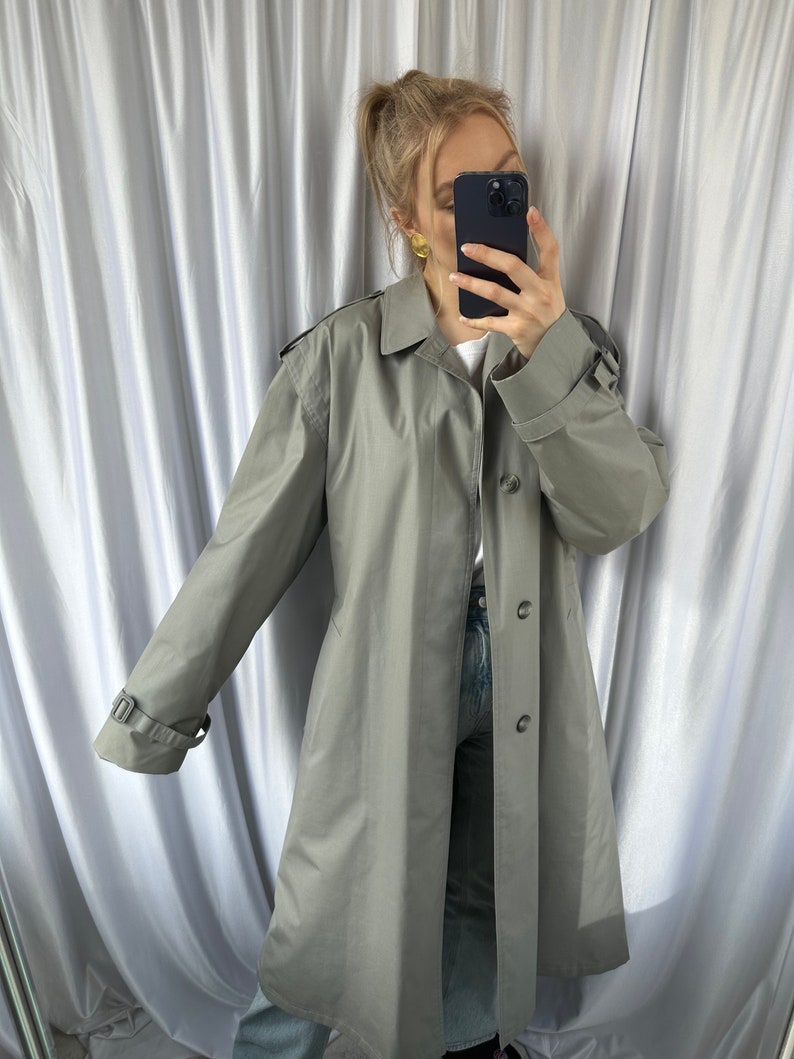 May include: A long, gray trench coat with a button closure and a belt. The coat has a classic design and is made of a lightweight fabric.
