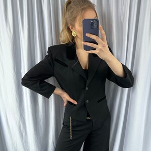 Może przedstawiać: Czarny blazer z aksamitnym kołnierzem i złotym łańcuszkiem. Blazer jest w połączeniu z czarnymi spodniami.