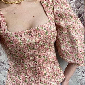 May include: A pink floral print blouse with a square neckline and puffed sleeves. The blouse has a button-up front and a fitted bodice.