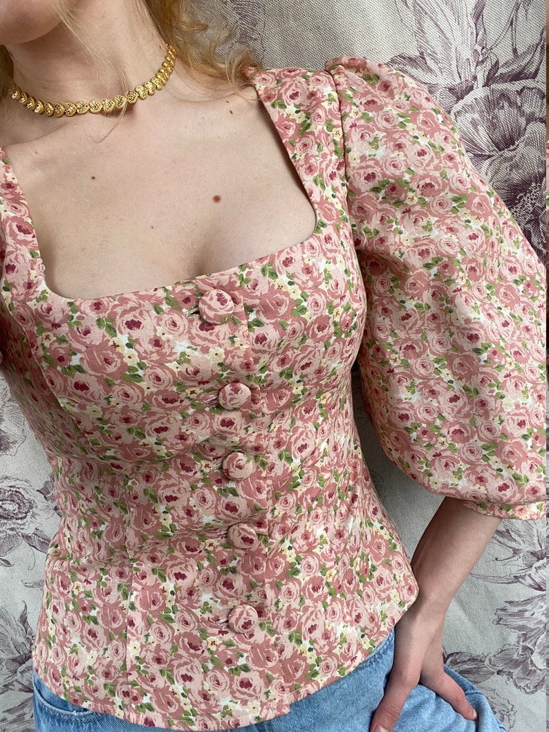 May include: A pink floral print blouse with a square neckline and puffed sleeves. The blouse has a button-up front and a fitted bodice.