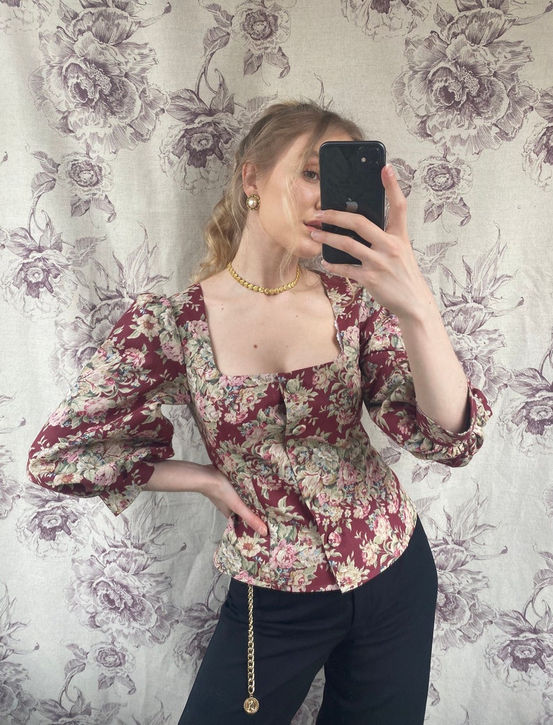 May include: A woman wearing a red and floral print blouse with a square neckline and puffed sleeves. She is also wearing black pants with a gold chain belt.