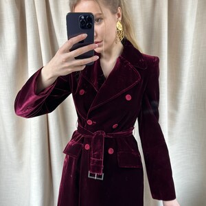 May include: A long, burgundy velvet coat with a double-breasted closure, a belt, and two pockets. The coat has a classic, tailored silhouette.
