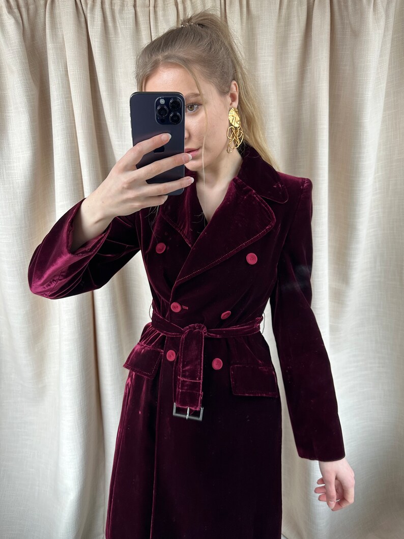 May include: A long, burgundy velvet coat with a double-breasted closure, a belt, and two pockets. The coat has a classic, tailored silhouette.