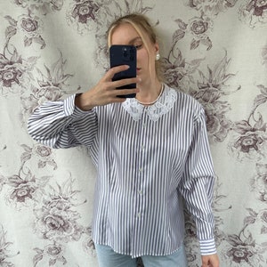 Pode incluir: Uma camisa de manga comprida listrada em branco e roxo com uma gola de renda branca. A camisa tem um fecho de bot&otilde;es na frente e mangas compridas com punhos de bot&otilde;es.