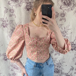 May include: A pink floral cropped top with puffed sleeves and a square neckline. The top has a button-up front and is made of a lightweight fabric.