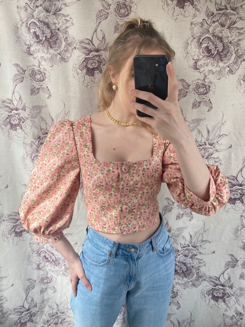 May include: A pink floral cropped top with puffed sleeves and a square neckline. The top has a button-up front and is made of a lightweight fabric.