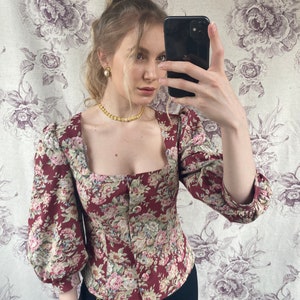 May include: A red and floral print blouse with a square neckline and long sleeves. The blouse has a button closure in the front and a fitted silhouette.