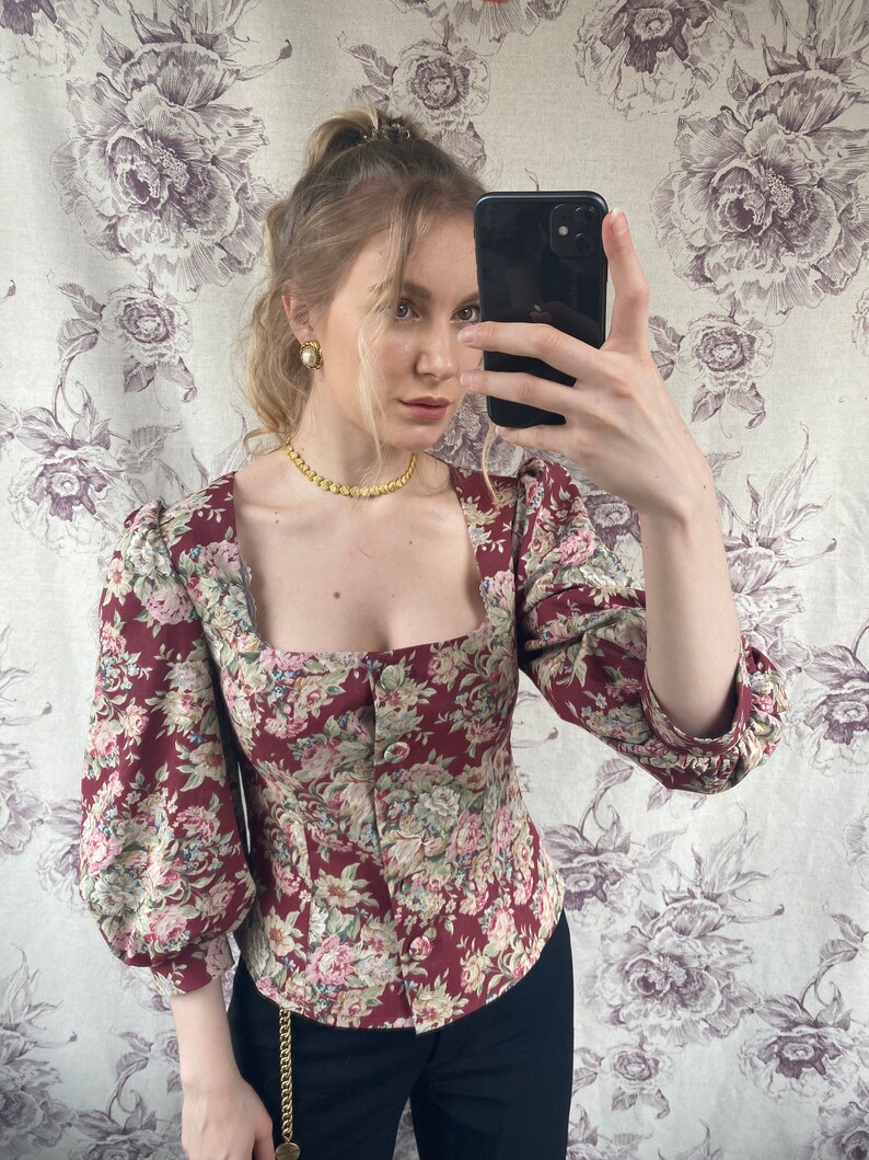 May include: A red and floral print blouse with a square neckline and long sleeves. The blouse has a button closure in the front and a fitted silhouette.