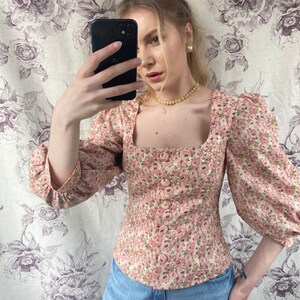 May include: A pink floral print blouse with a square neckline and puffed sleeves. The blouse is fitted and has a cinched waist.
