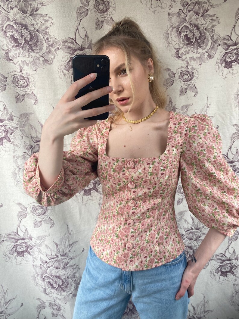 May include: A pink floral print blouse with a square neckline and puffed sleeves. The blouse is fitted and has a cinched waist.