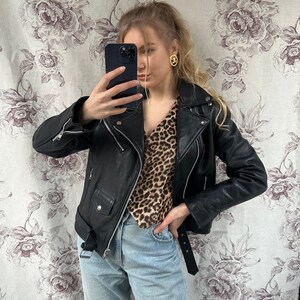 May include: A woman wearing a black leather jacket over a brown and black leopard print blouse. She is wearing light blue jeans and a black belt. She is holding a black smartphone in her right hand and taking a selfie.