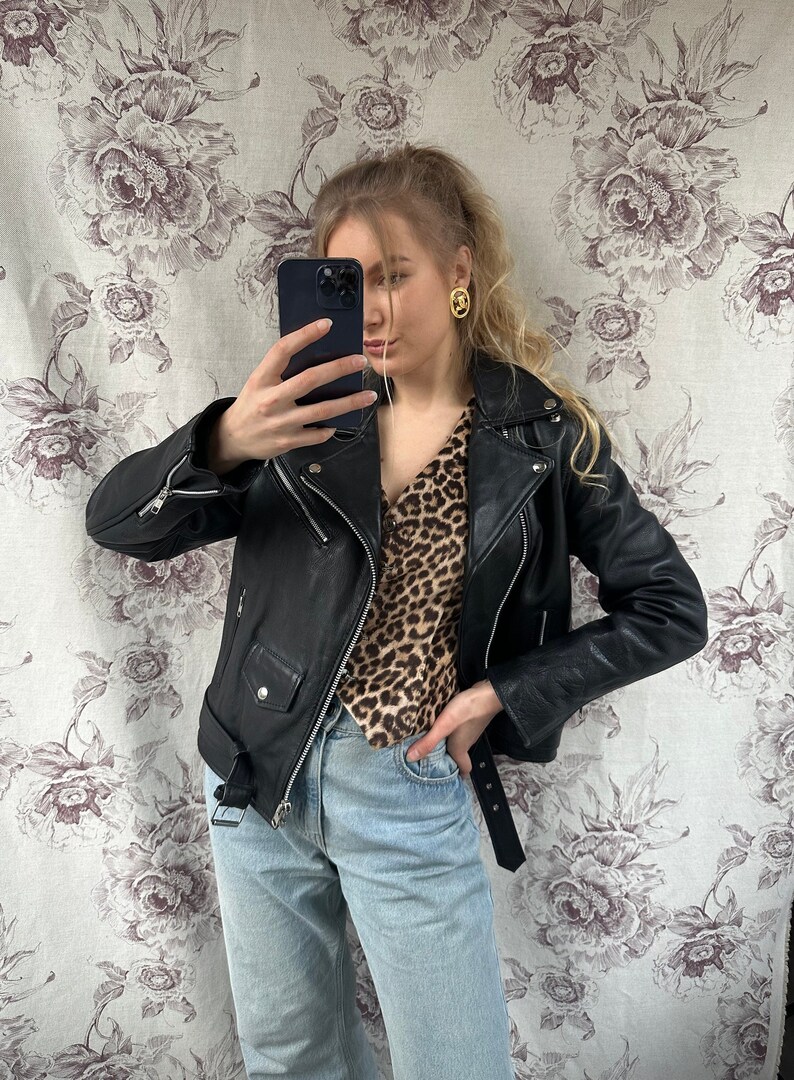 May include: A woman wearing a black leather jacket over a brown and black leopard print blouse. She is wearing light blue jeans and a black belt. She is holding a black smartphone in her right hand and taking a selfie.