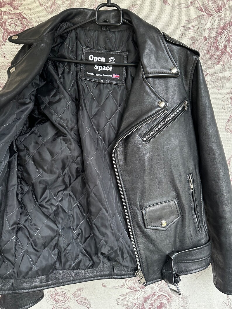 May include: Black leather motorcycle jacket with a quilted lining. The jacket has a zipper closure, two pockets, and a belt. The label on the jacket reads "Open Space" and "Quality Leather Garments".