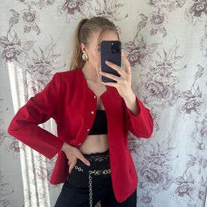 Puede incluir: Una chaqueta roja con botones dorados y un cintur&oacute;n de cadena dorada. La chaqueta se lleva sobre un top negro sin mangas y pantalones negros.