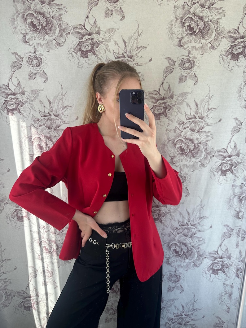 Puede incluir: Una chaqueta roja con botones dorados y un cintur&oacute;n de cadena dorada. La chaqueta se lleva sobre un top negro sin mangas y pantalones negros.