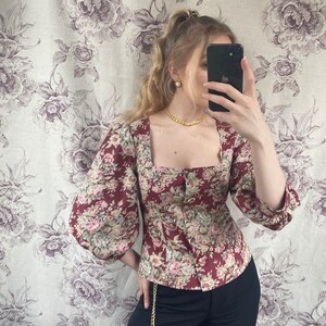 May include: A red and white floral print blouse with a square neckline and puffed sleeves. The blouse has a button-up front and is cinched at the waist. The model is wearing black pants with a gold chain belt.
