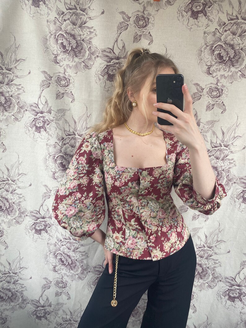 May include: A red and white floral print blouse with a square neckline and puffed sleeves. The blouse has a button-up front and is cinched at the waist. The model is wearing black pants with a gold chain belt.