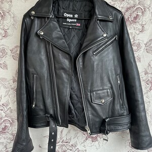 May include: Black leather motorcycle jacket with a zipper closure and a belt at the waist. The jacket has a label that reads "Open Air Space" and "Quality Leather Garments".