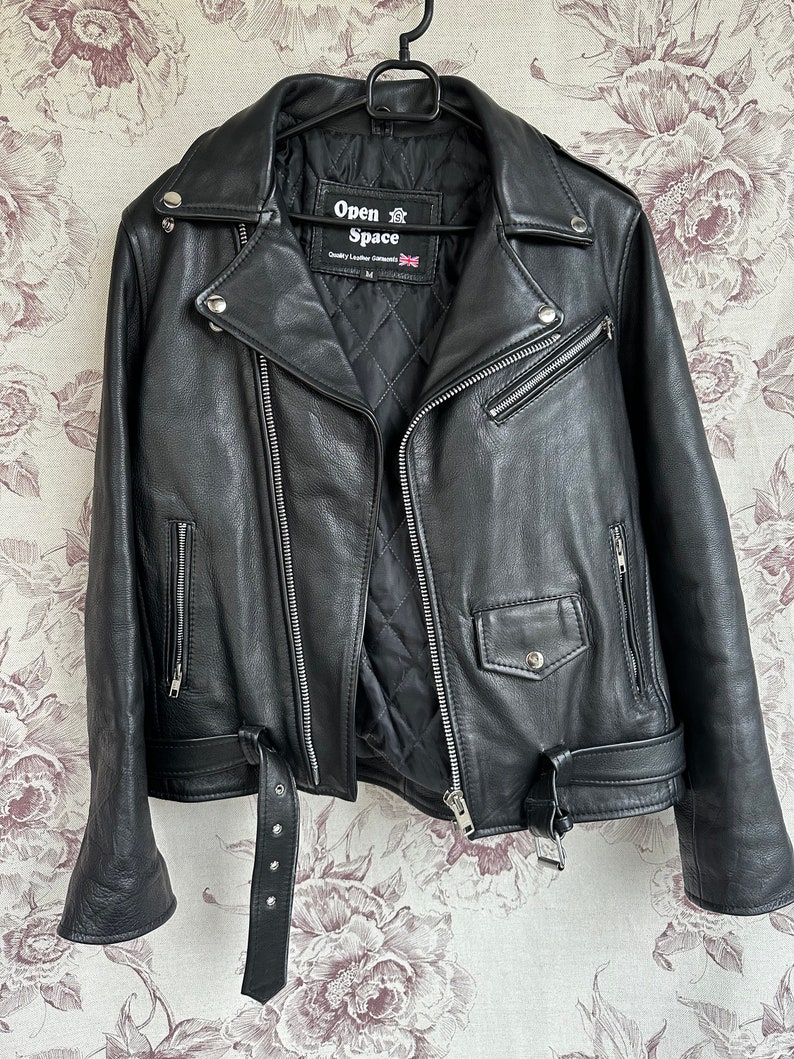 May include: Black leather motorcycle jacket with a zipper closure and a belt at the waist. The jacket has a label that reads "Open Air Space" and "Quality Leather Garments".
