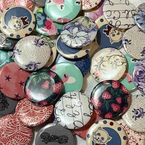 May include: A collection of round, colorful buttons with various patterns and designs. The buttons feature floral patterns, polka dots, stripes, and abstract designs. Some buttons have a white background with black text, while others have a black background with white text. The buttons are all approximately the same size and are arranged in a random pattern.