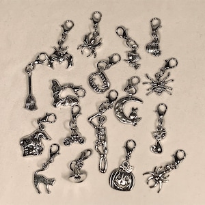 May include: A collection of 18 silver-toned Halloween-themed charms. The charms feature various spooky designs, including bats, spiders, pumpkins, skeletons, cats, and a witch's broom.