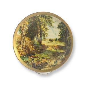 May include: A round, gold-rimmed trinket box with a painted scene of a rural landscape. The scene features trees, a stream, a path, and a flock of sheep.