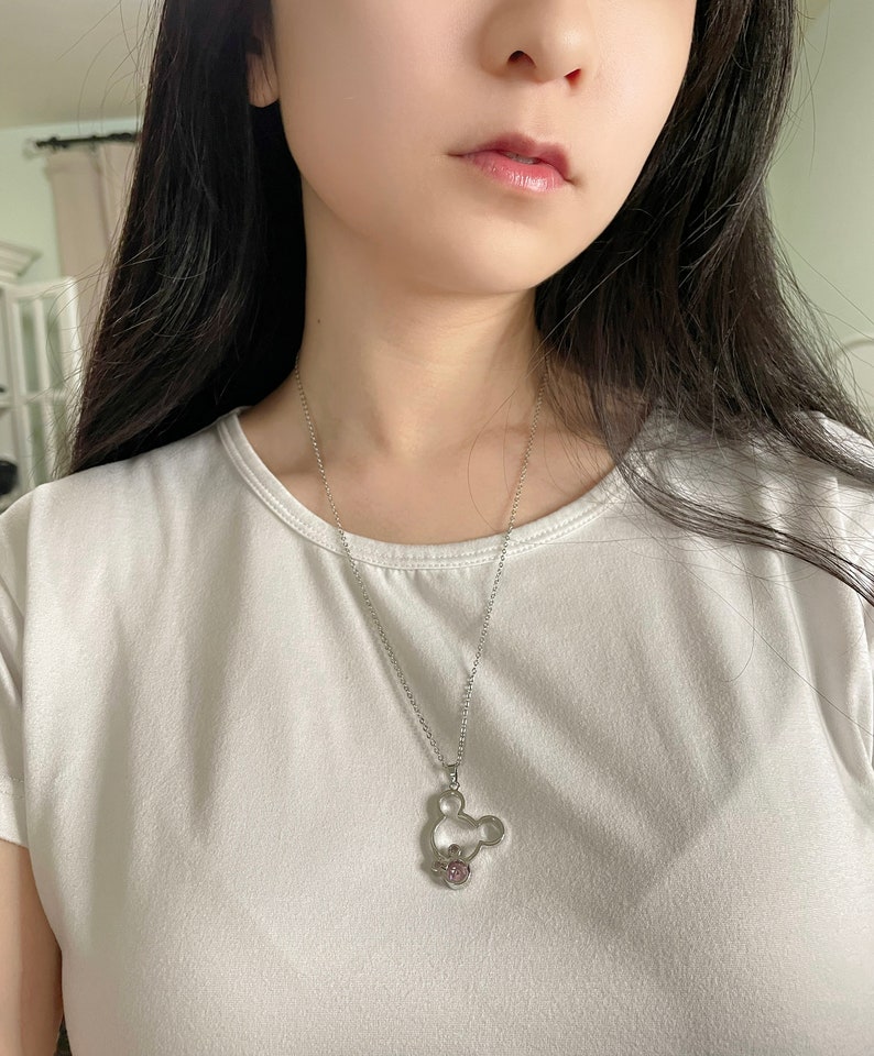 May include: A silver chain necklace with a Mickey Mouse-shaped pendant featuring a pink crystal. The pendant is hollow and has a delicate design.