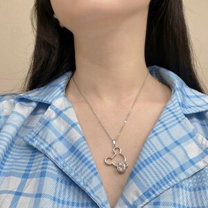 May include: A silver necklace with a Mickey Mouse pendant featuring a pink rhinestone. The necklace is worn by a person wearing a blue and white plaid shirt.