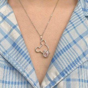 May include: A silver necklace with a Mickey Mouse pendant featuring a pink crystal. The pendant is a cutout of Mickey Mouse's head with a pink crystal in the center.
