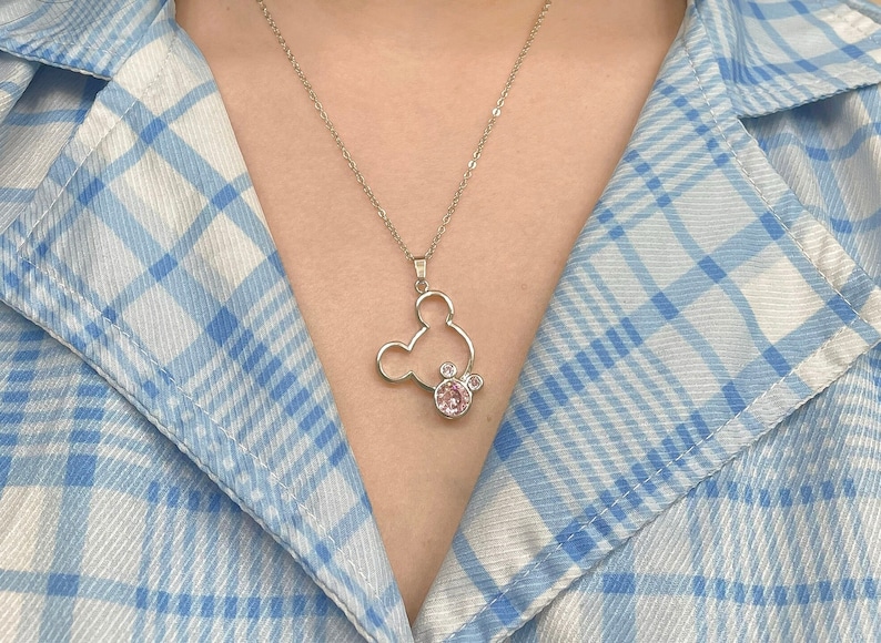May include: A silver necklace with a Mickey Mouse pendant featuring a pink crystal. The pendant is a cutout of Mickey Mouse's head with a pink crystal in the center.