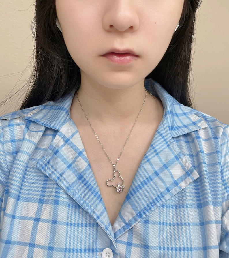 May include: A silver necklace with a Mickey Mouse pendant featuring pink rhinestones. The necklace is worn by a person wearing a blue and white plaid shirt.