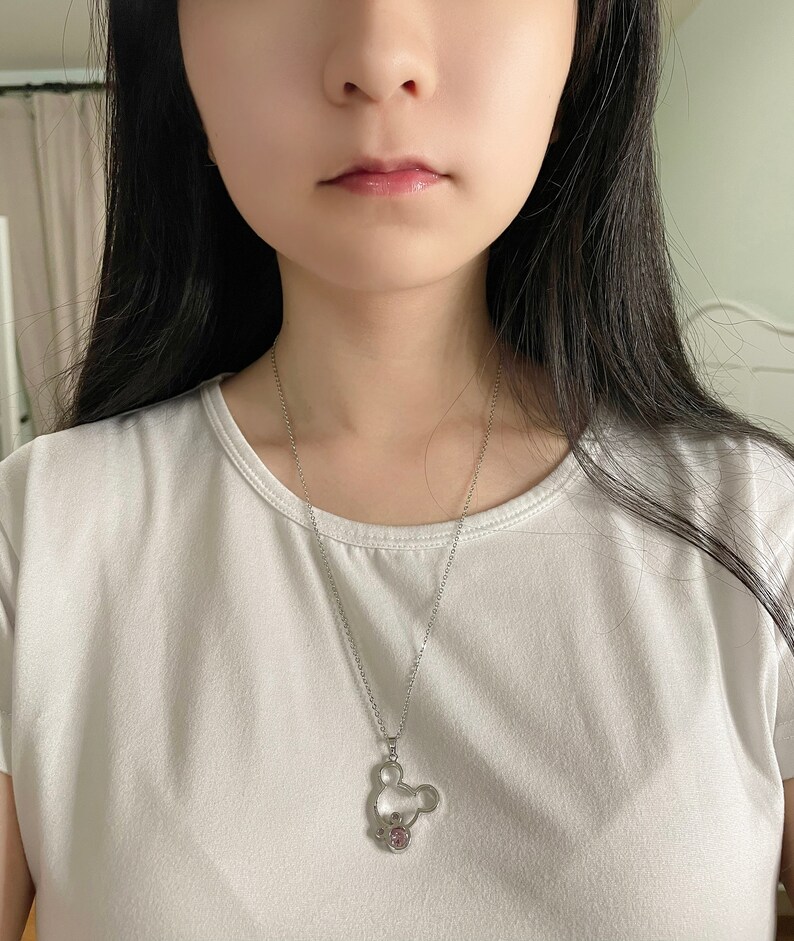 May include: A silver chain necklace with a Mickey Mouse pendant featuring a pink crystal. The pendant is a hollow outline of Mickey Mouse's head with a small pink crystal in the center.