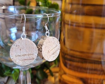 May include: A pair of cork earrings with silver hooks. The earrings are round and have a natural cork finish. The earrings are displayed on a wine glass with a bottle of wine in the background.