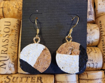May include: A pair of handmade earrings featuring circular cork pendants with gold-colored hooks. Each pendant is divided into three sections: white, brown, and black. The earrings are displayed against a backdrop of wine corks.