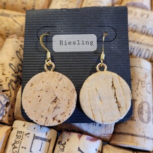 May include: A pair of cork earrings with gold hooks. The earrings are round and have a natural cork finish. The earrings are displayed on a black card with the word "Riesling" printed on it.