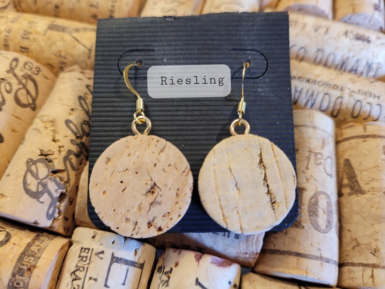 May include: A pair of cork earrings with gold hooks. The earrings are round and have a natural cork finish. The earrings are displayed on a black card with the word "Riesling" printed on it.