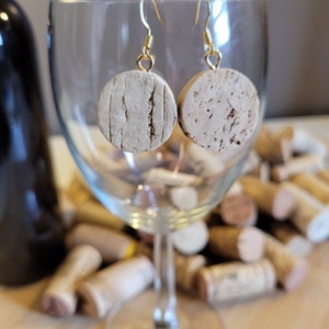 May include: A pair of cork earrings with gold hooks. The earrings are round and have a natural cork finish. The earrings are hanging from a wine glass.
