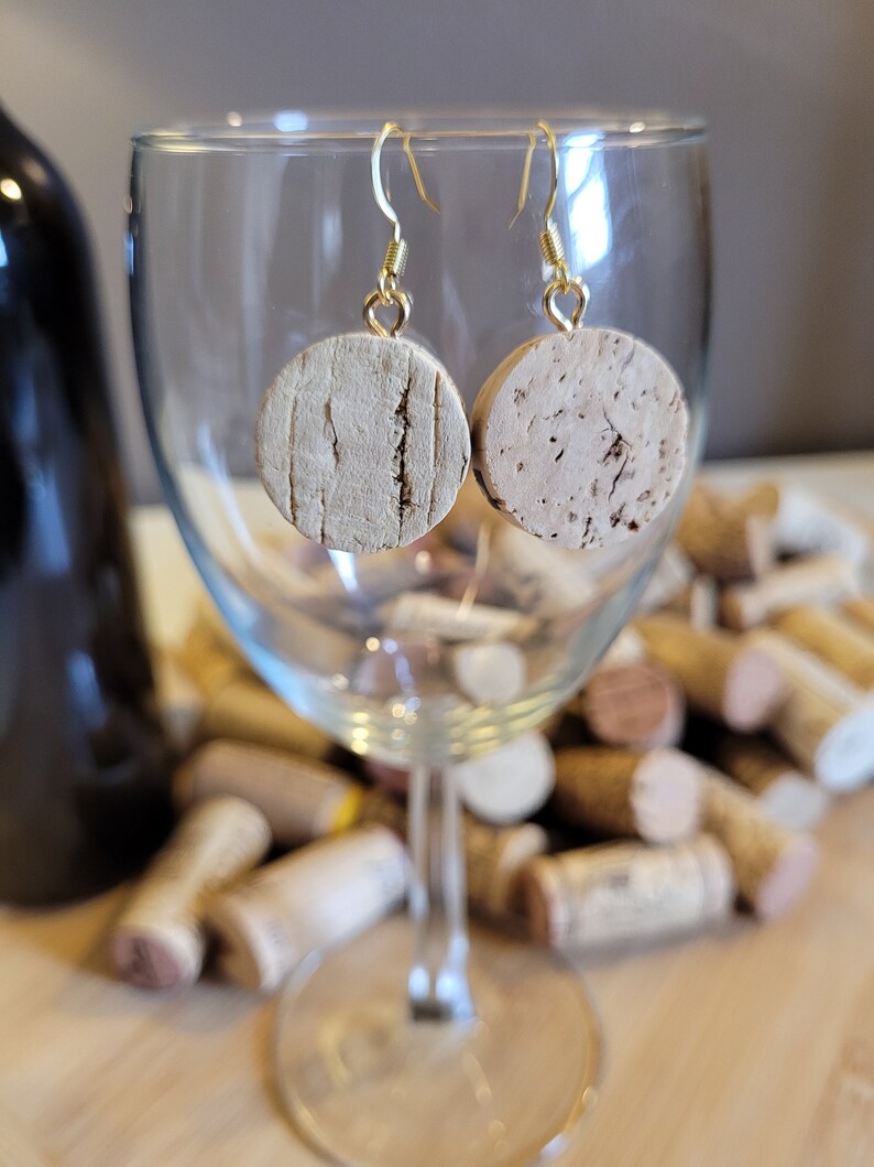 May include: A pair of cork earrings with gold hooks. The earrings are round and have a natural cork finish. The earrings are hanging from a wine glass.