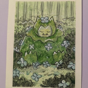 May include: Watercolor painting of a green creature with closed eyes, surrounded by blue flowers and foliage. The background features a forest scene with vertical tree trunks. The artwork is signed "Savannah Davenport".