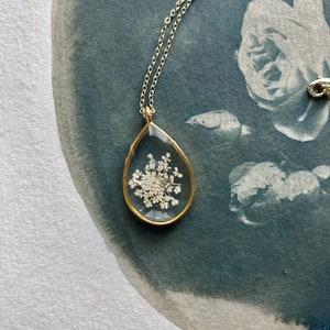 Könnte beinhalten: Eine zarte goldene Halskette mit einem tropfenförmigen Anhänger. Der Anhänger enthält eine gepresste weiße Blume, die einem kleinen Strauß ähnelt.