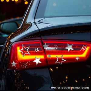 May include: A black car rear taillight assembly featuring a series of silver star-shaped decals applied to the red and yellow lens surface. The stickers vary in size and include both solid and outlined star designs across the light housing.
