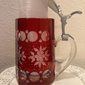 May include: A ruby red glass beer stein with a clear glass handle and a metal hinged lid. The stein features white floral and circular etched designs. The stein is cylindrical with an octagonal base.
