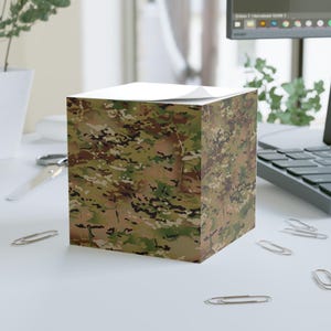 May include: A square stack of adhesive note paper featuring a multi-colored camouflage pattern in shades of olive green, brown, tan, and black. The paper block has a flat top surface and vertical sides displaying the printed military-style design.